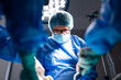 © Graphicroyalty - Focused senior male surgeon wearing glasses, blue scrubs, surgical mask and cap performs operation under bright LED light in hospital operating room.