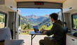 © Hira - Young man working on laptop at table inside camper van with open back doors overlooking beautiful mountain landscape