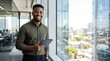 © Eve - Smiling young black businessman holding tablet by office window confident male entrepreneur professional corporate worker successful manager portrait modern workspace commercial concept