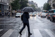 © KATSIARYNA - Person crossing a city street under an umbrella during heavy rain, illustrating urban weather, commuting, and daily life.