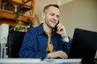 © DragonImages - Young adult Caucasian man speaking on smartphone and typing on laptop in office, handling customer support and communication. Ideal for call center, remote work, business service
