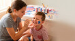 © Kristelinmind - Teenage girl painting blue star on cheek of a little boy for Independence Day celebration. Sibling fun with arts and craft at home. Patriotic holiday activity for America commemoration.