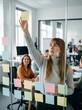 © Yurgentum - A young woman in a meeting room arranges colorful sticky notes on glass, fostering collaboration.