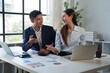 © Tj - Two Asian business professionals actively collaborating, discussing financial data, and analyzing reports on laptops and papers during a productive meeting in a bright office environment