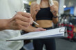 © Natee Meepian - Personal trainer discussing workout plan with client on clipboard in modern gym for fitness coaching