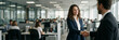 © Aleksei Solovev - A smiling woman in a blue blazer shakes hands with a man in a dark suit in a bright office setting with coworkers in the background.