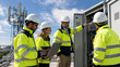 © Kowit - Four technicians in high-visibility jackets and helmets inspect and maintain telecommunications equipment outdoors under a partly cloudy sky.