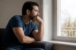 © 3AN - Sad mid adult man sitting on sofa looking out window with thoughtful expression. Lonely guy in living room feeling depressed or stressed at home.