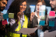 © ultramansk - Smiling Black female engineer writes ideas on a sticky-note glass board while three diverse team members observe in a dark collaborative workspace.