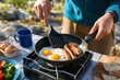 © Studio - Fried eggs and sausages cooking in frying pan on portable camping stove outdoors during sunny morning adventure trip, close-up