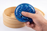 Hand squeezing a glittery blue dumpling squishy inside a bamboo steamer box on a white background