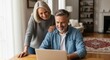 © Dara - A smiling elderly couple sits at a wooden desk, working on a laptop together. The woman gestures towards the screen while the man smiles and leans on her shoulder.