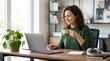 © Jing - Smiling woman working on laptop computer at home office desk, drinking coffee, enjoying remote work lifestyle with houseplant and bookshelf background.