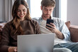 © luciano - Young couple on a sofa, woman typing on a laptop and man checking his smartphone