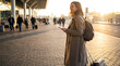 © Anthichada - A smiling woman with a backpack and suitcase holds a passport outside an airport terminal at sunset