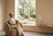 © Kassiopeia  - Senior woman sits in a warm beige room, looking out at a garden with trees during a sunny day in a quiet setting