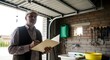 © pika - Man in cap and vest holding a folder and pen, standing in a garage with tools and a sink.