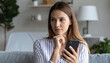 © SolaruS - A thoughtful young woman sits on a cozy sofa, holding her smartphone and looking away dreamily, lost in pleasant thoughts or planning her day in a bright, modern living room.