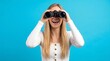 © Yassine - Happy young blonde woman looking through black binoculars against a vibrant solid light blue background studio