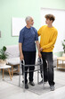 © New Africa - Caregiver. Teenage boy helping elderly man with walking frame indoors