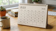 © Ibnu Fatah - July 2026 desk calendar stands on a wooden table next to a coffee mug, books, and plants in a cozy office setting.
