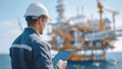 © Kingdola - Oil Rig Engineer: An engineer wearing a hard hat and safety vest stands on an oil rig, holding a tablet and looking out at the ocean. The rig is in the background, with a clear blue sky above.