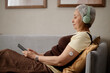 © TrueFrame Collective - Senior Asian woman wearing glucose sensor, listening through headphones and using tablet on sofa, managing digital health routine. Ideal for diabetes care, telehealth, wellness marketing