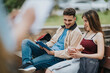 © qunica.com - Two students sit on a bench studying and laughing while sharing a book. A candid moment of young friends relaxing outdoors and using a phone between study sessions.