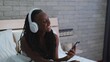 © ekaterinabyuksel - Cute dark-skinned woman listening to music on headphones while sitting in her bed, relaxing at home in her bedroom