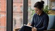 © LooPanda-Pictures - Focused Mindset: A young woman sits in a chair by a large window, deeply engrossed in her work on a tablet. Natural light illuminates her focused expression as she writes.
