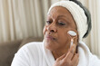 © Wavebreak Media - Senior African American woman sitting on sofa in white robe and terry headband using facial roller