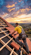 © alchena - Professional roofer installing red clay tiles on a house roof at sunset. Male builder in yellow shirt working on home construction with beautiful orange sky and scenic hills background.