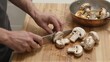 © joeycheung - Person slicing mushrooms on wooden board near copper pan with sauteed pieces.