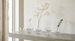 © Soklin - A minimalist arrangement of clear glass vases and a single branch of dried flowers on a concrete table in front of a window with sheer curtains.