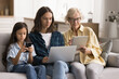 © fizkes - Multiple generations family, older granny, her grown-up daughter and pre-teen granddaughter using diverse gadgets seated on couch, looking engrossed and focused. Screen time awareness, tech overuse