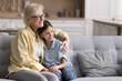 © fizkes - Loving older granny hugging her pretty little granddaughter seated on comfortable sofa looking into distance with positive, optimistic expression, daydreaming, spend priceless time together at home