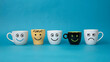 © Ali - A row of five ceramic mugs with different funny faces on a blue background
