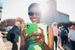 © BullRun - Joyful black female tourist holding mobile phone with both hands against sunny urban background. Cheerful African American model using online navigation or banking application in city center.