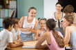 © JackF - Young boys and girls of different ages sit at table, playing strategy board game with colorful moving pieces while young woman teacher supervises