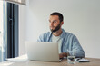 © Daniel - Young man with a beard looking away, reflecting on ideas or problems while remote working at a desk with a laptop, finding inspiration and concentration in a bright home office setting