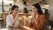 © mihrzn - Two smiling young women relax and chat while sipping refreshing green smoothies at a wooden table inside a bright, modern cafe with warm lighting.