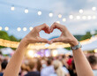 © T2flair - A person forms a heart shape with their hands against a blurred background of twinkling lights and a crowd, symbolizing love and connection.