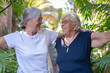 © luciano - Two cheerful senior adult women smiling and embracing each other, enjoying a moment of connection and friendship in a vibrant green tropical park