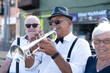 © luciano - Senior man playing a trumpet while wearing a fedora, bowtie, and suspenders, entertaining an appreciative senior couple smiling in the background