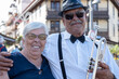 © luciano - Senior man musician in a fedora and bow tie holding a trumpet, standing with his happy mature adult woman partner, both enjoying urban street life and a journey together