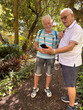 © luciano - Two senior adult men sharing a moment of outdoor leisure and friendship, looking at a smartphone together while walking through a lush botanical garden on vacation
