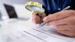 © Mahu - Businessman examining documents with magnifying glass, office desk with laptop, close-up hands holding pen and loupe