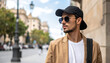 © baa - Man wearing sunglasses and a baseball cap, looking away with a serious expression, standing on a city street with blurred buildings and trees in the background, urban fashion, modern style