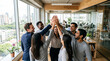 © Tofayel Ahmed - Energetic diverse team of six stands in a group high-five celebration inside a modern timber-lined office with large windows overlooking a green city skyline, great for team success content.