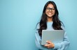© miss irine - Young Indian woman with glasses smiles holding laptop. She wears blue sweater. Confident student ready for work or study. Person works on computer against blue background.